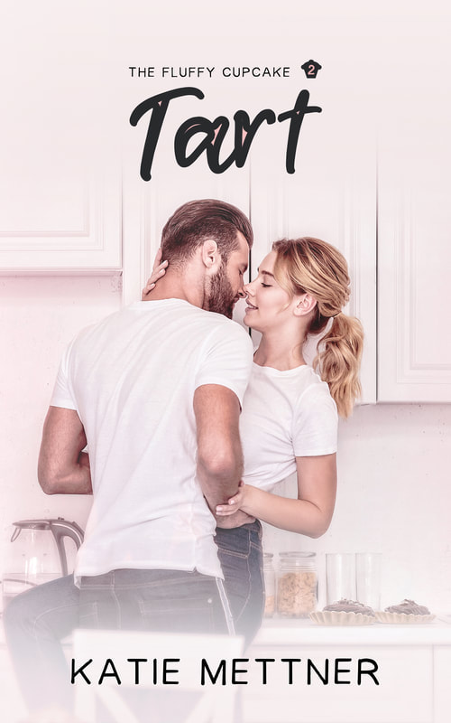 The background is a kitchen and there is a couple standing in the middle of it. They are close together, almost kissing. He is wearing a white t-shirt and jeans. She is also wearing a white t-shirt and jeans. She has a long blonde ponytail and he has short brown hair and a beard. At the top it says, The Fluffy Cupcake 2 Tart, Katie Mettner
